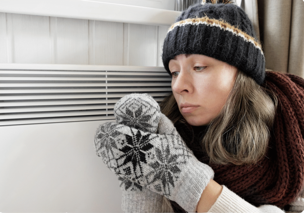 Woman dressed in winter clothes trying to warm up near a broken radiator in a UK home